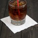 A glass of brown liquid with ice and a straw on a white beverage napkin.