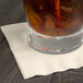A glass of liquid with ice and a straw on an ivory Creative Converting beverage napkin.