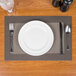 A Taos beige Snap Drape PVC placemat on a table with a white plate, fork, and spoon.