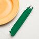 A fork and knife wrapped in an emerald green Creative Converting paper dinner napkin on a table next to a plate.
