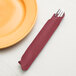 A fork and knife in a burgundy Creative Converting paper napkin next to a plate.