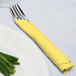 A fork and knife wrapped in a yellow Creative Converting luncheon napkin on a table next to a plate of green onions.