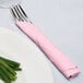 A fork and knife wrapped in a pink Creative Converting Classic Pink napkin next to a plate of green onions.
