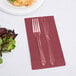 A fork and knife on a burgundy Creative Converting guest towel next to a plate of salad.