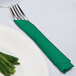 A fork and knife wrapped in an emerald green Creative Converting luncheon napkin on a plate.