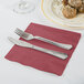 A burgundy 1/4 fold luncheon napkin with a fork and knife on a table next to a plate of food.
