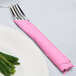 A fork and knife wrapped in a pink Creative Converting luncheon napkin next to a plate of green onions.