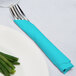 A fork and knife wrapped in a blue Bermuda Blue napkin on a plate.