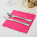 A Creative Converting hot magenta pink luncheon napkin with a fork and knife on a pink surface.