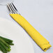A fork and knife wrapped in a yellow Creative Converting 3-ply luncheon napkin on a plate with green onions.