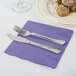 A silver fork and knife on a purple Creative Converting luncheon napkin next to a plate of food.
