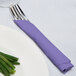 A fork and knife wrapped in a purple Creative Converting luncheon napkin on a white plate.