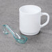 A white mug and a clear green Fineline Tiny Tensils plastic spoon on a grey surface.