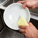 A hand using a 3M Scotch-Brite Dobie All Purpose Cleaning Pad to wash a plate.