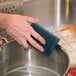 A hand holding a 3M Scotch-Brite Extra Heavy-Duty Pot 'n Pan Handler Scour Pad over a metal bowl of water.