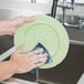 A hand using a 3M Scotch-Brite blue power pad to wash a green plate.