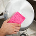 A person's hand holding a pink 3M Scotch-Brite sponge over a pan.