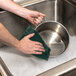 A person washing a metal pan with a 3M Scotch-Brite green scouring pad.
