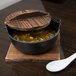 A Thunder Group cast iron Japanese noodle bowl with a wooden lid on a table with a white spoon.