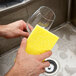 A hand using a 3M Scotch-Brite light-duty yellow sponge to wash a wine glass over a sink.