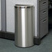 A silver Commercial Zone Precision stainless steel half round trash receptacle next to a desk.