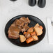 A Hall China Foundry black coupe plate with meat and vegetables on it.
