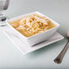 A Libbey Ultra Bright White square porcelain bowl filled with soup and noodles with a spoon.