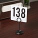An American Metalcraft black swirl base card holder with a white sign and black numbers on a wooden table.