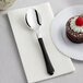 A Visions heavy weight plastic spoon with a black handle on a napkin next to a dessert.