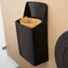 A black Lavex wall-mount sanitary napkin receptacle with a brown paper bag inside.