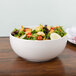A white Arcoroc porcelain bowl filled with salad with tomatoes, lettuce, croutons, and other vegetables.