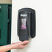 A person installing a black GOJO True Fit wall plate for a hand sanitizer dispenser on a wall.