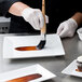 A gloved hand uses an Ateco round pastry brush to paint brown sauce on a piece of cake.