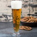 A close up of a GET Tritan plastic footed pilsner glass full of beer with a bowl of pretzels on a table.