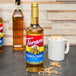 A Torani Bourbon Caramel flavoring syrup bottle next to a mug of foamy drink on a wood table.