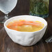 A bowl of 10 Strawberry Street bright white porcelain pinch soup with carrots and noodles next to a glass of water.
