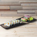 A rectangular stoneware slab holding sushi rolls on a wood surface.