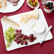 A white rectangular wood grain porcelain platter with cheese, grapes, and a round white cheese with a cut out slice.