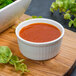 A white porcelain fluted ramekin filled with red sauce on a cutting board.