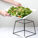 A person holding a plate of salad on an American Metalcraft black square display stand.