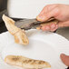 A person's hand holding Vollrath stainless steel tongs with a tan handle over a plate of chicken.