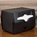 A Vollrath black tabletop napkin dispenser with black and white napkins on a wooden table.