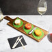 A Tablecraft acacia wood paddle serving board with food on it next to glasses of white wine.