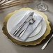 A white charger plate with a scalloped gold rim with silverware on a napkin.