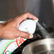A hand holding a white Pan Sprease spray can over a silver pan on a kitchen counter.