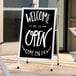 An Aarco satin aluminum A-frame sign with white text that says "Welcome Open Come In" on a sidewalk.