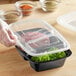 A gloved hand holding a Choice black plastic container with 2 compartments filled with food.