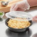 A person in gloves holding a Choice black plastic container with macaroni and cheese and a plastic lid.