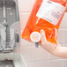 A hand holding a plastic bag of orange liquid with a grey GOJO Spa Bath dispenser