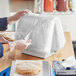 A person in gloves and a white apron using a Choice Deli Saddle bag stand to put food in a plain plastic bag.